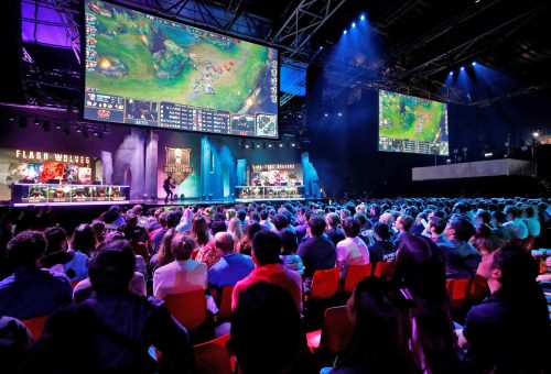 In a stadium, fans spectate a League of Legends esports tournament on a large widescreen TV